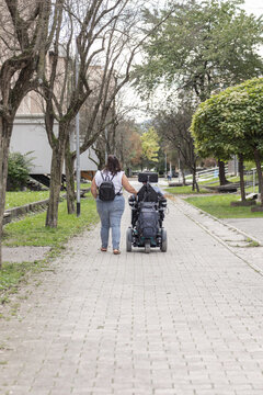 A man in a wheelchair is being assisted by a woman on a city sidewalk. Another person walks nearby.