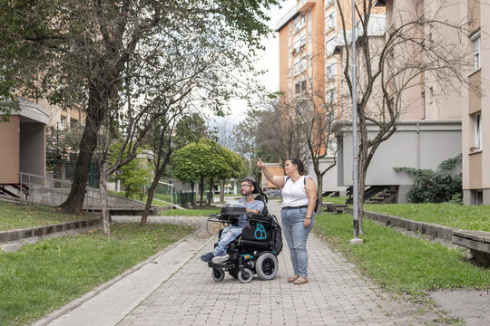A man in a wheelchair is on a path while a woman helps him. They are in a city with buildings and trees around them.