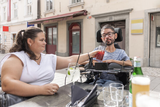 A woman helps a man in a wheelchair enjoy a drink at a restaurant. They are seated outside on a social outing.