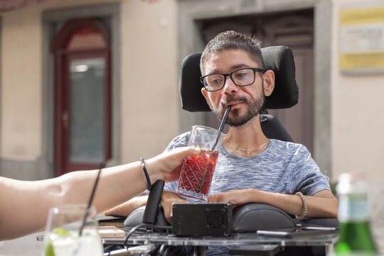 A man in a wheelchair drinks from a glass straw while sitting outside a restaurant. His personal assistant helps him with holding drink.