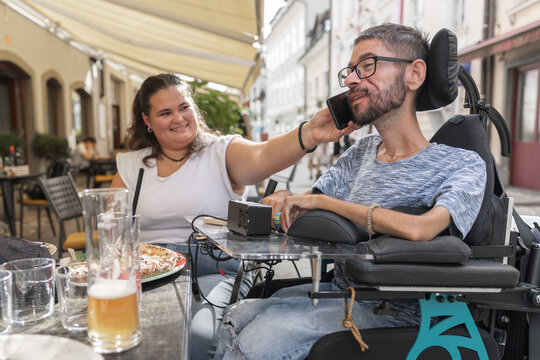 A man in a wheelchair is dining with a woman at a restaurant. They are enjoying food and drinks while talking on mobile phone.