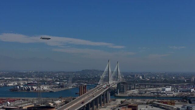 Aerial view of the Gerald Desmond Bridge spanning the Port of Long Beach with a blimp flying in the clear blue sky in Long Beach, California, United States.