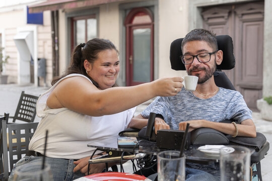 A woman assists a man in a wheelchair as they dine at a restaurant. They share food and coffee in an accessible setting.