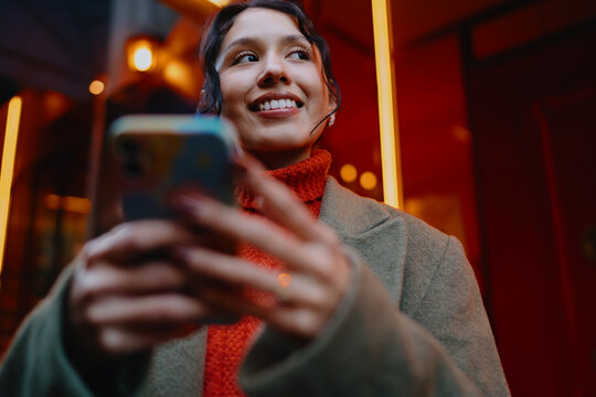 Young multiracial lady with smartphone looking away and smiling in city at night, mobile connectivity, digital lifestyle, 5G wireless network, startup culture, inclusion, urban traveler mood.