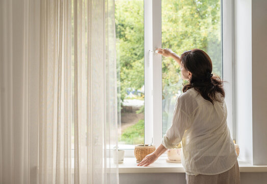 Mature woman stands by window and opens it, letting in fresh air and natural light, practice that promotes ventilation and nature-oriented lifestyle. Slow living, mindful lifestyle, healthy habits