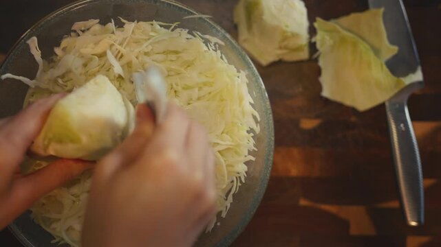 Top view close-up of woman's hands shredding fresh cabbage with a peeler into a full glass bowl on a wooden cutting board. Cooking and healthy food preparation concept in a home kitchen. 