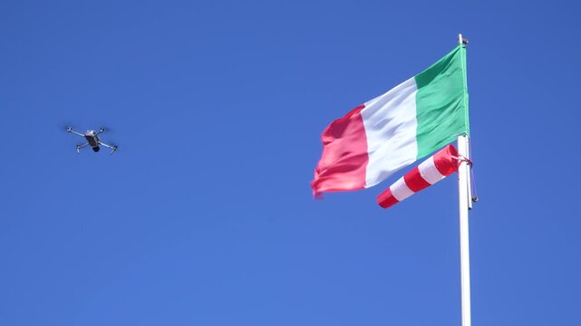 Drone facing strong wind, with an Italian flag and a windsock in the background