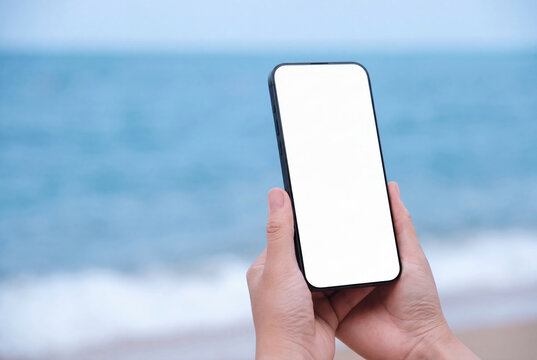 Smartphone mockup held by hands at the beach. Blank white screen display. Ocean waves and sandy shore background