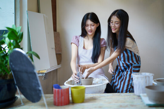 Two Young Asian Women wearing aprons collaborate or Learning Pottery on Spinning Wheel in Art Studio Workshop, art education and hobby concepts