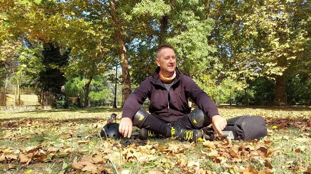Man in roller skates and knee protection sitting on grass with dry leaves in city park, taking a break after rollerblading on a sunny fall day. Commercial B-roll, 4K UHD