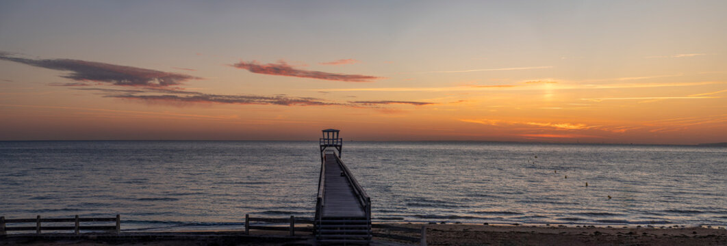Luc-Sur-Mer, France - 08 08 2026:  Panoramic view of a wooden pier in front of the beach, sea, a colorful sunrise and Le Havre city far away