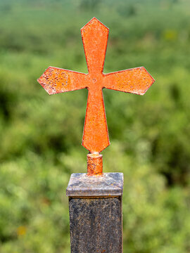 Eisernes orangefarbenes Kreuz auf rostigem Pfahl vor gr&uuml;ner Wildwiese