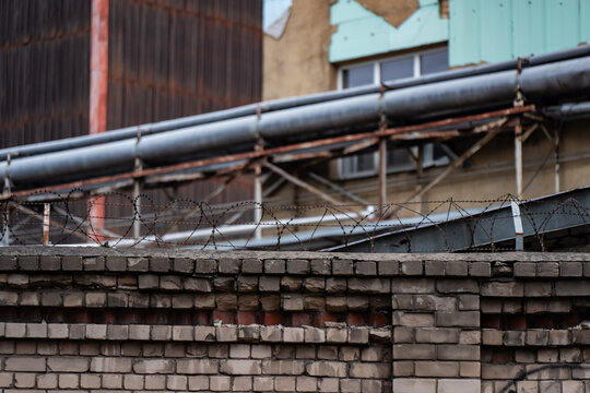 Coiled security wire tops a weathered brick boundary wall below industrial pipework, illustrating perimeter protection components and maintenance needs at an aging industrial site
