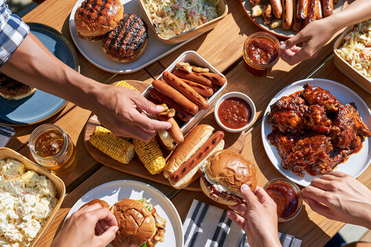 Hands reach for food at a vibrant picnic table. Buns, sausages, corn, and wings fill the spread. Laughter and casual energy fill the scene. Perfect for summer gatherings or food marketing
