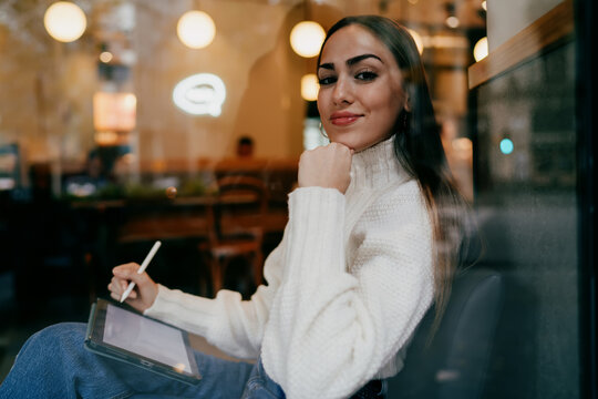 Professional female illustrator smiling at camera while working on digital tablet in cafe, creative freelancer holding stylus pen, modern remote work lifestyle, authentic representation.