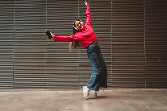 Cheerful girl jumping with joy listening to favorite tracks in headphones, young woman holding phone and dancing in urban setting, success and happiness concept, full length body portrait.