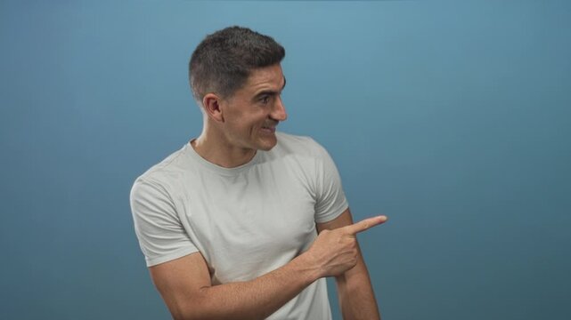 Man points finger to right in studio blue backdrop wearing light tee, looking to side and smiling; friendly invitation.