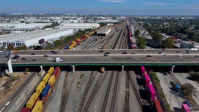 Aerial view of a highway overpass crossing a busy rail yard with freight trains and colorful shipping containers in Long Beach, California, United States.