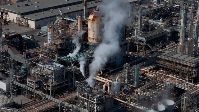 Aerial view of an industrial oil refinery with complex piping, metal structures, and steam rising from chimneys in Carson, California, United States.