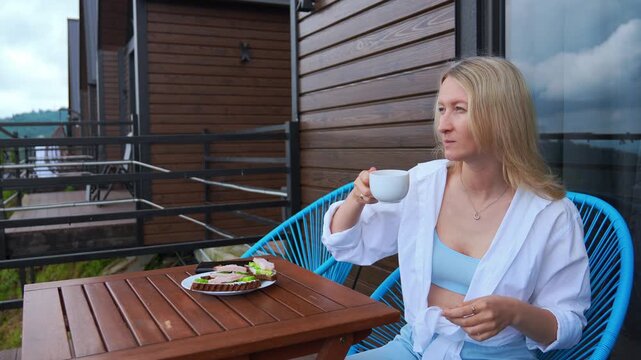 Blonde woman sitting at a wooden table on the balcony of a modern house, enjoying a sandwich and a hot drink for breakfast while appreciating the peaceful morning atmosphere of her holiday