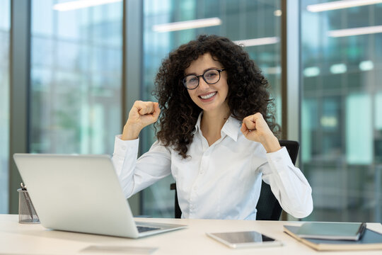 Happy businesswoman with curly hair and glasses celebrating triumph, raising fists in a gesture of victory while working on her laptop in a modern office environment