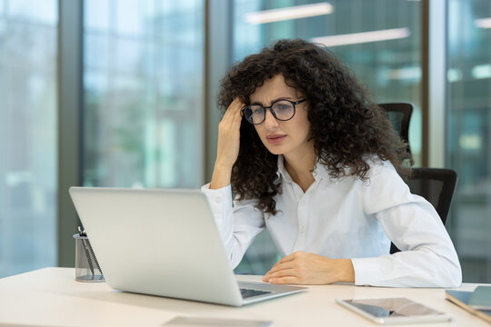 Stressed young business woman with curly hair and glasses holding her head, experiencing a headache and burnout while working on a laptop in a modern office
