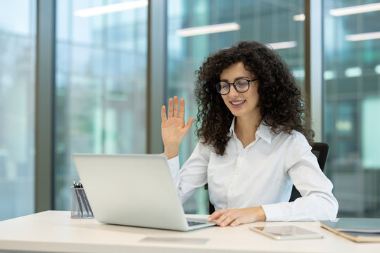 Young smiling businesswoman with curly hair and glasses waving hand during online video call on laptop, engaging in remote work from a contemporary office environment