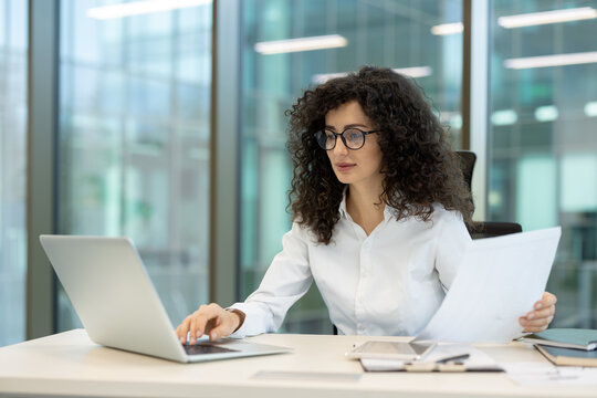 Young businesswoman with curly hair and glasses concentrating on her work, typing on a laptop and holding paperwork, representing business tasks and productivity in a modern office environment