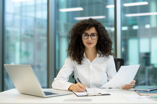 Businesswoman with curly hair and glasses reviewing paperwork and writing notes while working on a laptop at her modern office desk, focusing on productivity and professional tasks