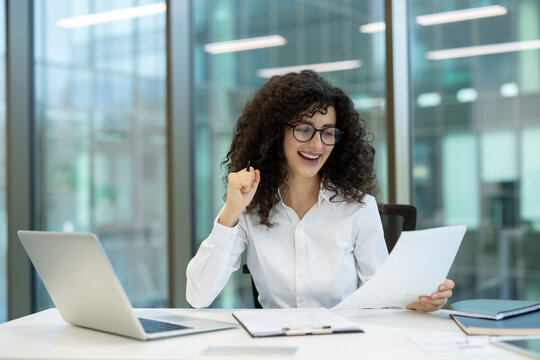 Young professional woman celebrating achievement with a fist pump while reading positive results on documents at her desk in a modern corporate office environment