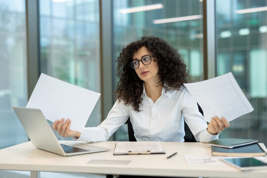 Businesswoman juggling multiple tasks and papers while working on a laptop at her office desk, facing professional pressure and feeling overwhelmed by her busy workflow