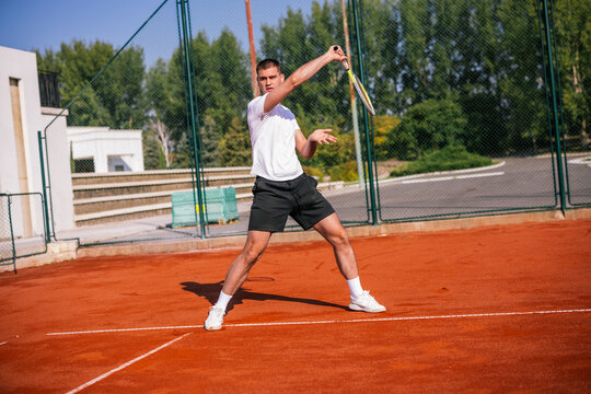 Young male tennis player performing high forehand volley swing on clay court.