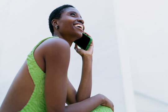 African American woman mobile call talking. Black female model using cellphone network roaming during trip, laughing while discussing international travel route near white wall copy space.
