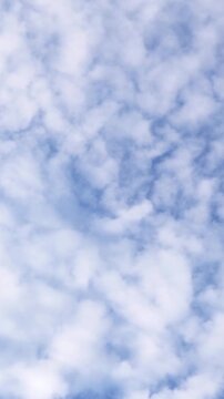 Vertical video looking up at soft, fluffy white cumulus clouds drifting across a bright blue sky.