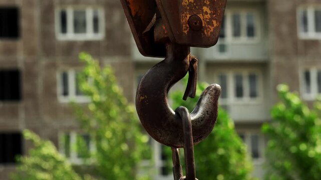 solid steel crane hook hangs suspended outdoors against backdrop of modern residential high rise. sturdy lifting hardware sways slightly in front of city apartment building and trees.