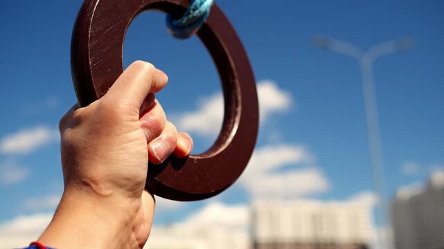 Hand holds gymnastics ring tightly against bright blue sky and distant city buildings. Athlete grips wooden training ring with determination during outdoor street workout on sunny day.