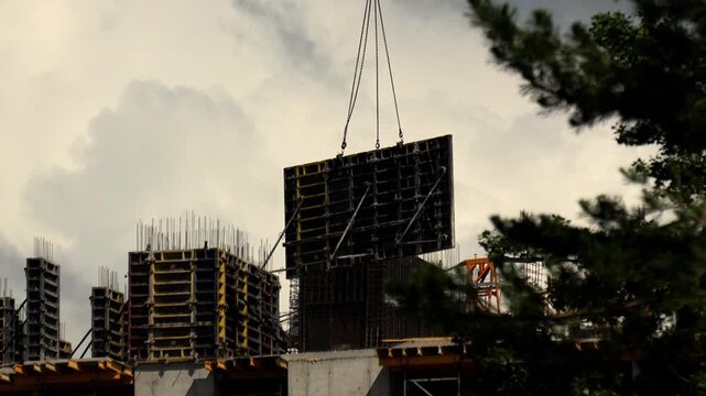 Formwork panel hangs from crane cables during heavy lifting operation on building structure. Industrial machinery positions structural component above reinforced concrete level at daytime.