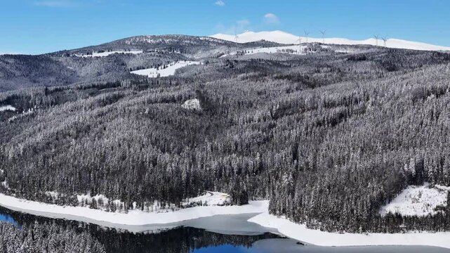 Aerial view of the snow-covered Soboth Reservoir surrounded by dense pine forests and distant mountains under a clear blue sky in Soboth Reservoir, Styria, Austria.