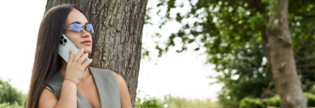 Young confident woman enjoying a sunny day while talking on her phone by a tree