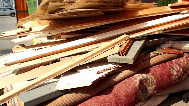 Stacked building timber and unwanted furnishings create large waste pile outdoors on sunny day. Demolition materials show worn wooden boards and old fabrics ready for collection and removal.