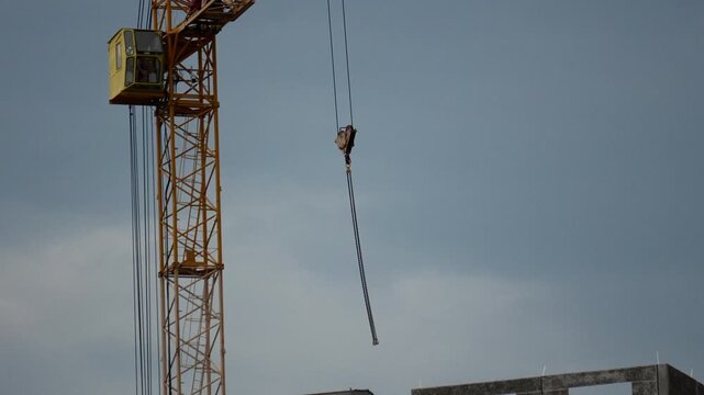 Tower crane rotates slowly across cloudy daytime sky at urban construction site. Yellow machine moves heavy metal cables toward concrete building structure during industrial development.