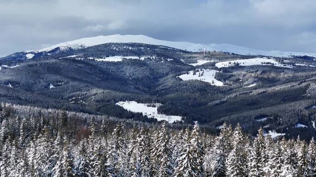 Aerial view of snowy mountains and pine forests with dense evergreen trees and rolling hills under a cloudy sky in Soboth Reservoir, Styria, Austria.
