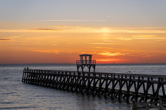 Luc-Sur-Mer, France - 08 08 2026:  Panoramic view of a wooden pier in front of the beach, sea, a colorful sunrise and Le Havre city far away
