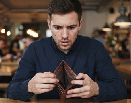 Young man sitting at a table holding an open empty wallet looking stressed about money