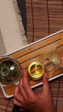 Top view of person hand holding glass pitcher and pouring hot tea into small glass cup on wooden tray. Authentic Chinese tea ceremony atmosphere with bamboo mat background.