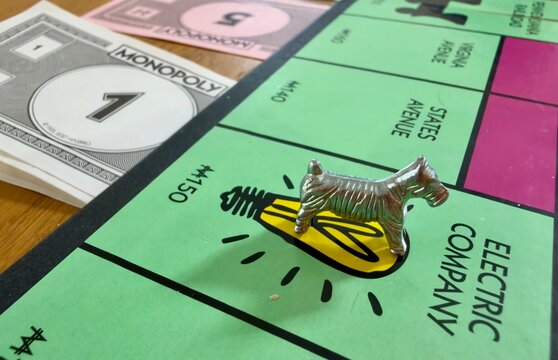Close-up shot of a classic Monopoly board game featuring the iconic metal Scottie dog playing piece resting on the green Electric Company property space. Stacks of play money including $1 and $5 bills