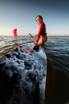 Woman holding kitesurfing board in water with kite in background