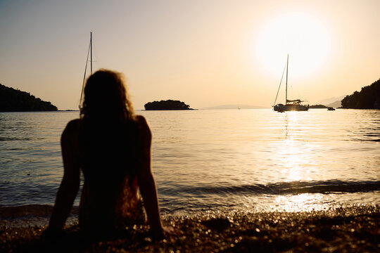 Silhouette Of Woman Sitting On A Beach Watching Sailboats At Golden Hour Sunset Over Calm Sea