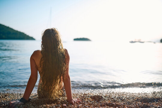 Woman Sitting on Pebble Beach Facing Calm Sea at Sunset Relaxing on Summer Vacation