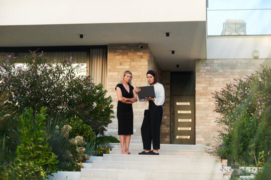 Real Estate Meeting Outside Modern Home: Female Agent Showing Property Details to Client on Laptop